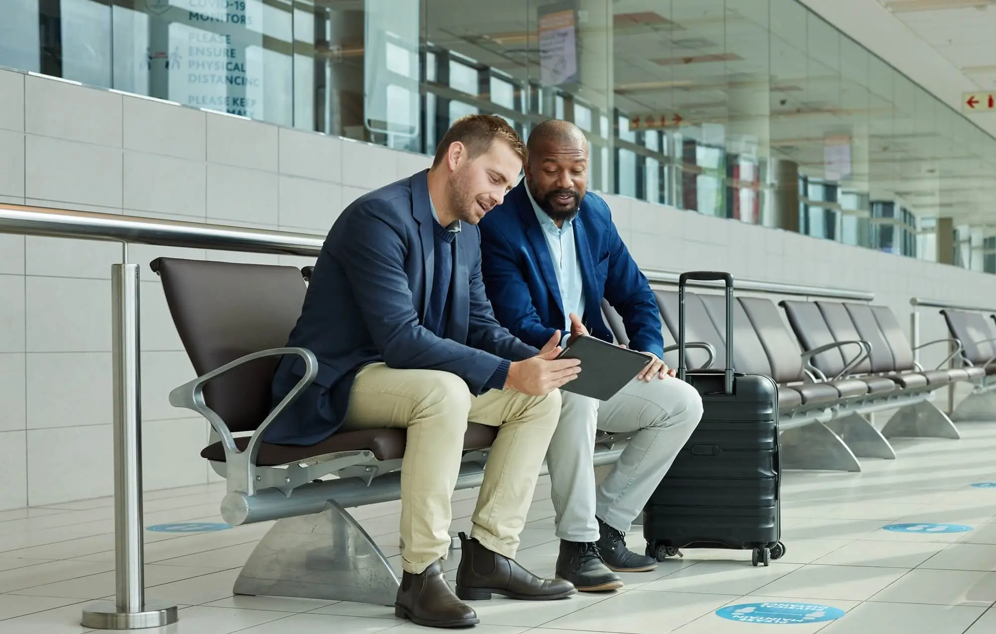 Two men in business attire seated in an airport terminal looking at a tablet with luggage beside them