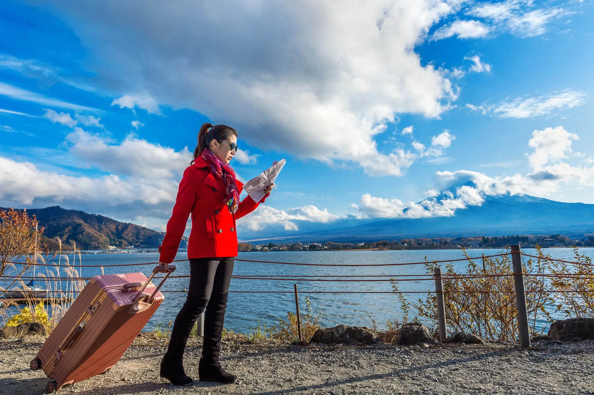 A traveler in a red coat pulling a suitcase while checking a map beside a scenic waterfront, representing the start of the holiday travel season