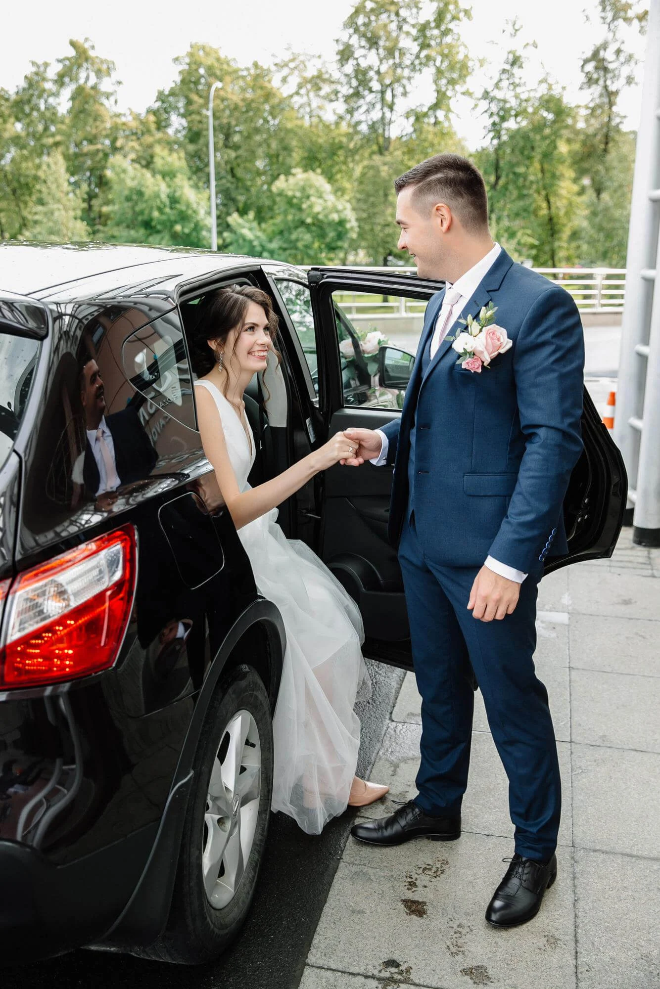 Bride exiting a car, helped by a groom in a blue suit with a floral boutonniere.