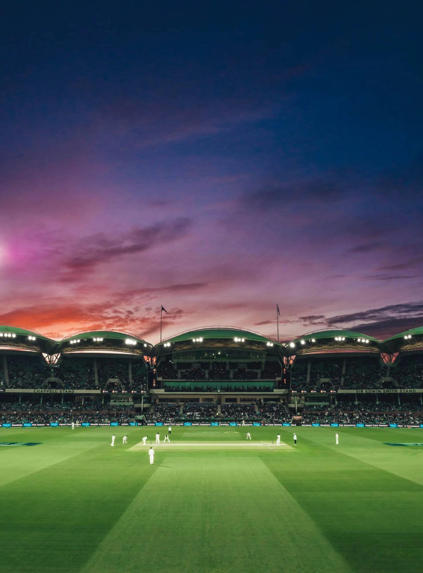 Stadium with a cricket match at sunset, under a colorful sky.