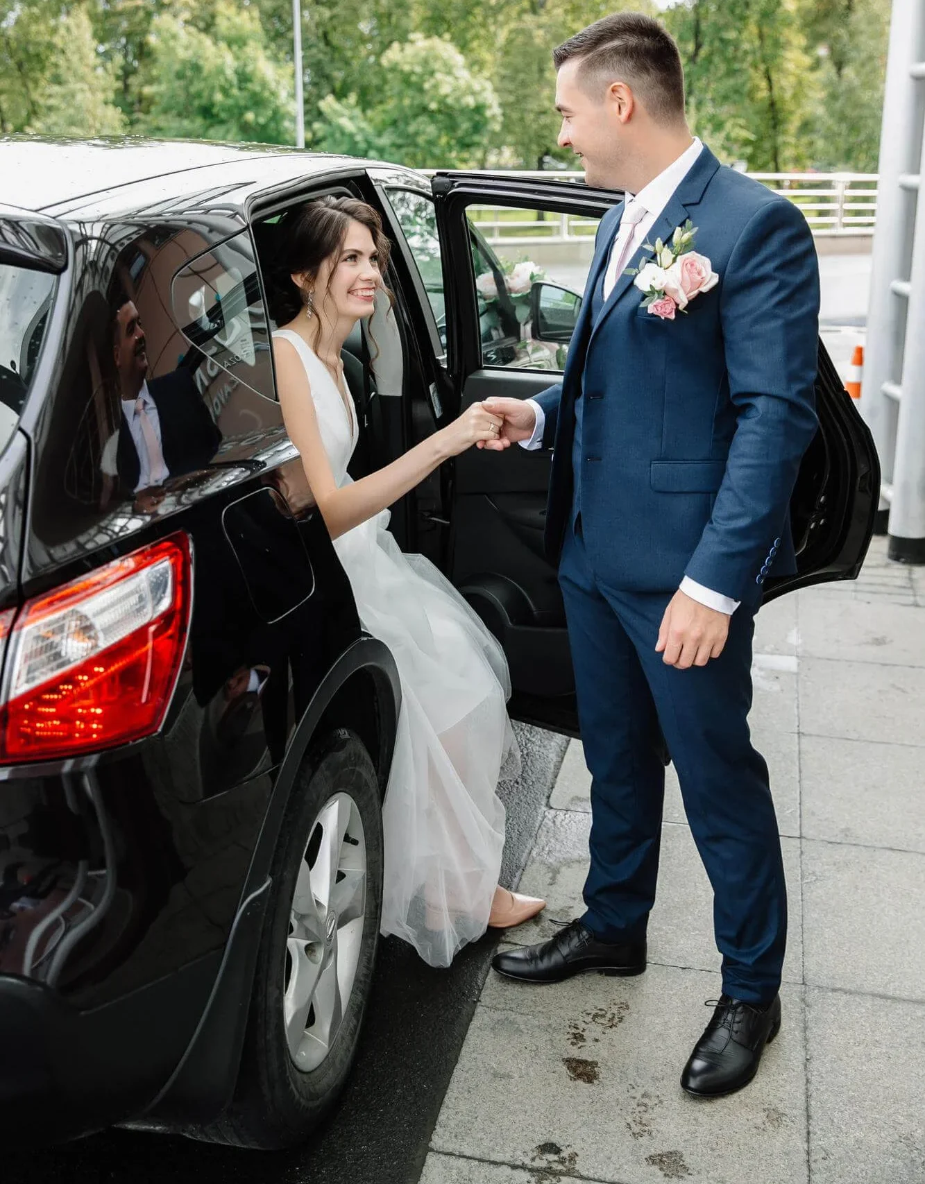 A man in a suit helps a woman in a wedding dress out of a car.