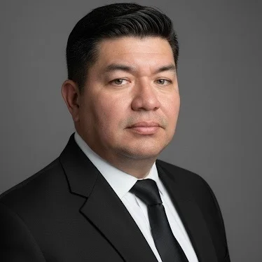 A man in a black suit and tie stands against a plain gray background.