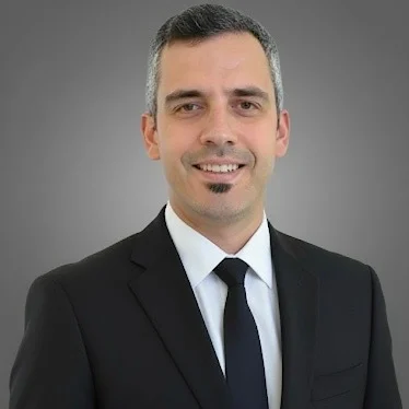 A smiling man in a suit stands against a gray background, possibly representing VOY Black Car's experienced team.