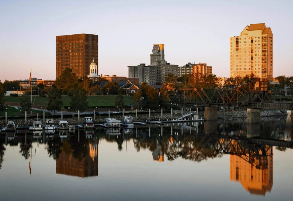 City skyline reflecting on a calm river at sunset, with boats docked at the waterfront.