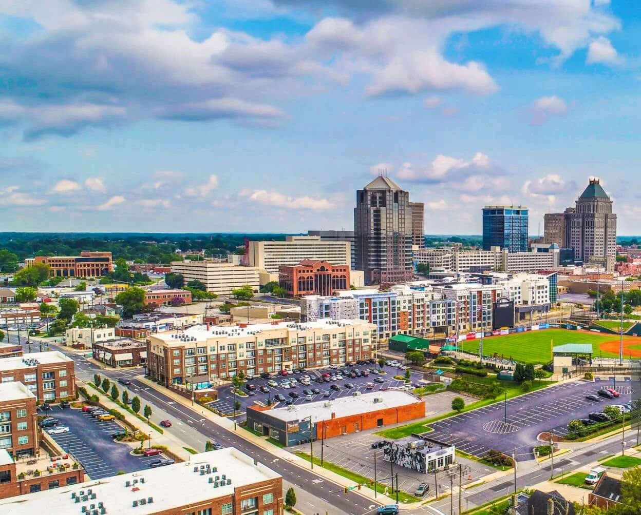 Aerial view of Greensboro's skyline, featuring tall buildings, residential areas, and a sports field under a partly cloudy sky.