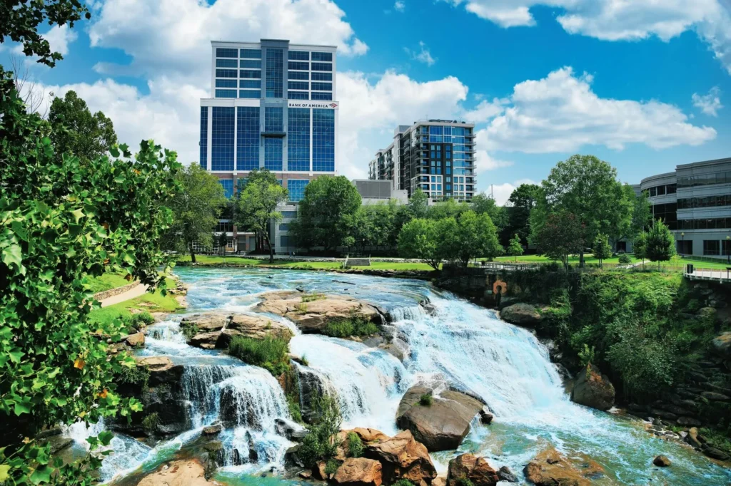 Cityscape with a waterfall in the foreground, surrounded by greenery, under a blue sky with scattered clouds.