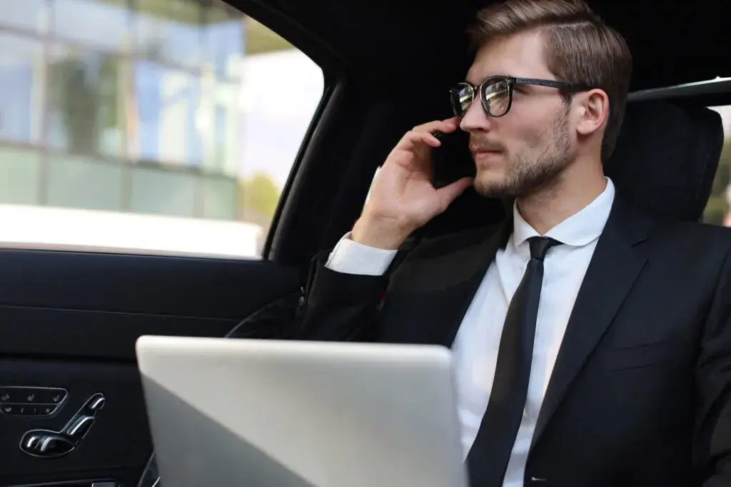 Man in a suit using a smartphone and laptop inside a car.