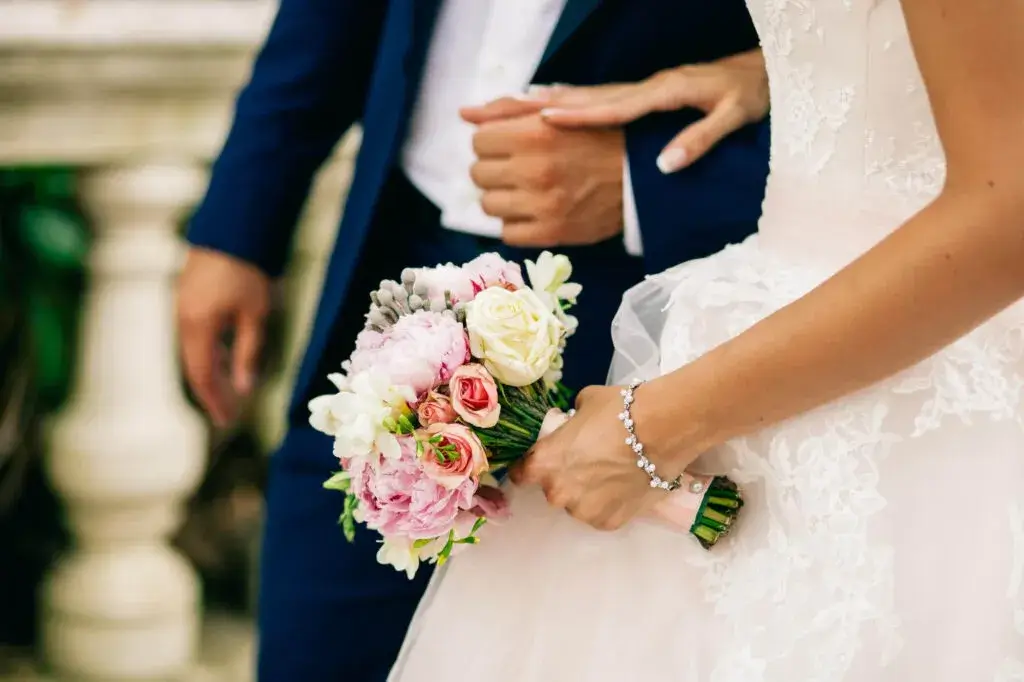 Bride holding bouquet with pink and white flowers, arm linked with groom in a suit.